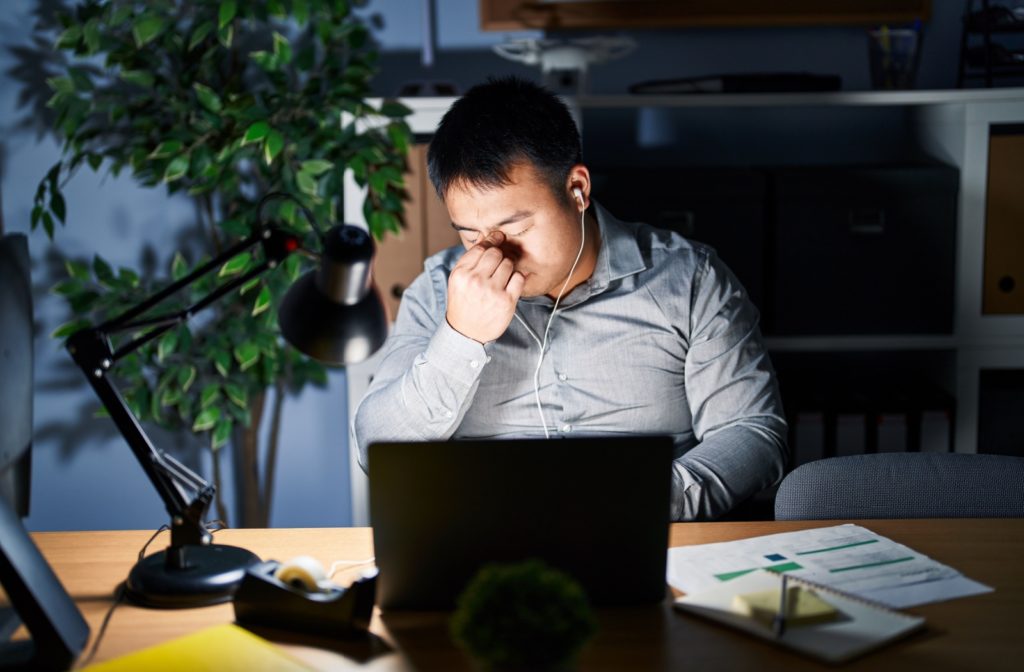 A man rubbing his eyes while working in a a dimly lit room.