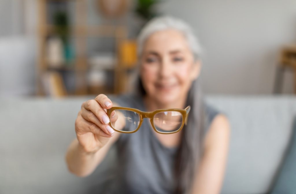 An out of focus older woman holds out her reading glasses