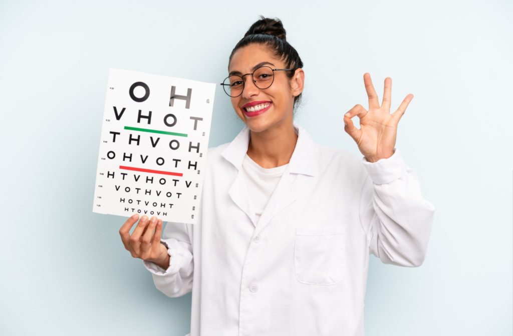 A happy optometrist holds up an eye chart and gives the gesture for okay.