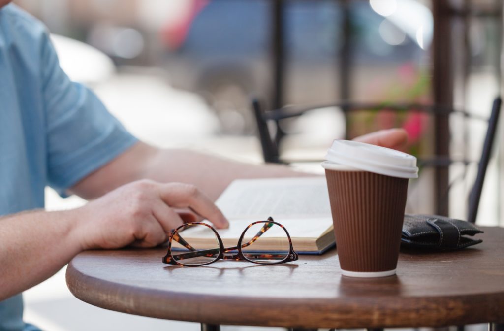 A man reading a book at an outdoor table, his eyeglasses set aside along with a coffee cup