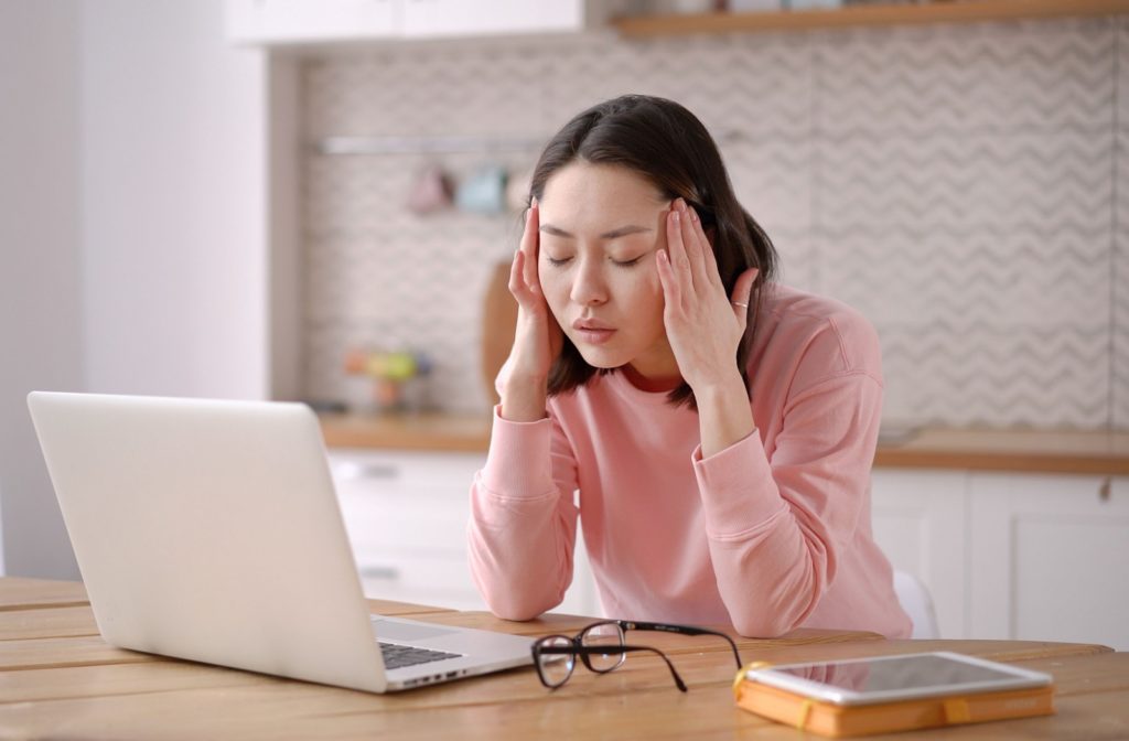 A woman in a pink sweater massages a headache at her temples, her eyeglasses cast aside on the table as she uses her computer