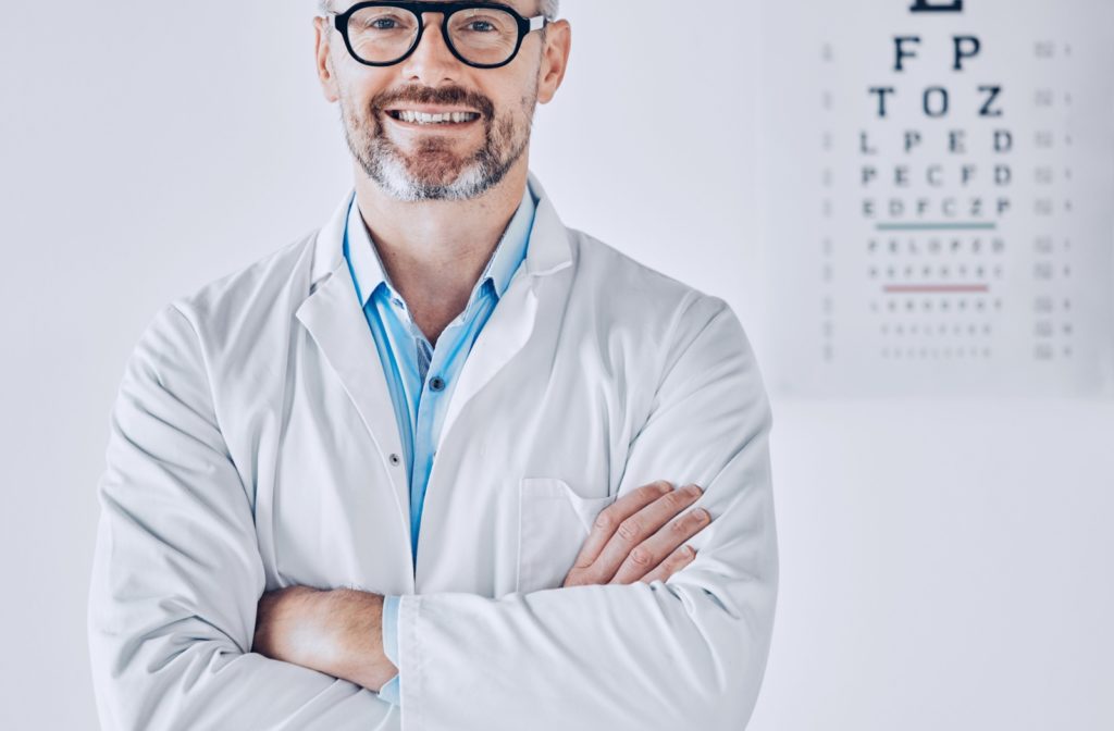 An image of a smiling optometrist with their arms crossed and a Snellen chart as a backdrop.