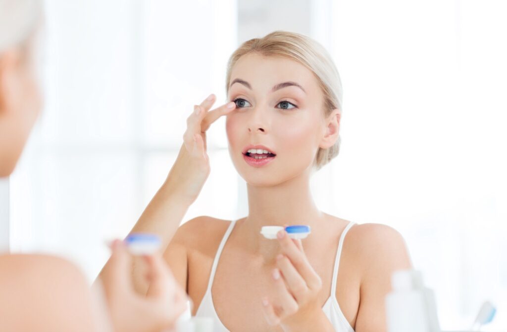A person standing in front of a mirror in a brightly lit room using their index finger on their right hand to insert a contact lens while holding a contact lens case with their left hand.