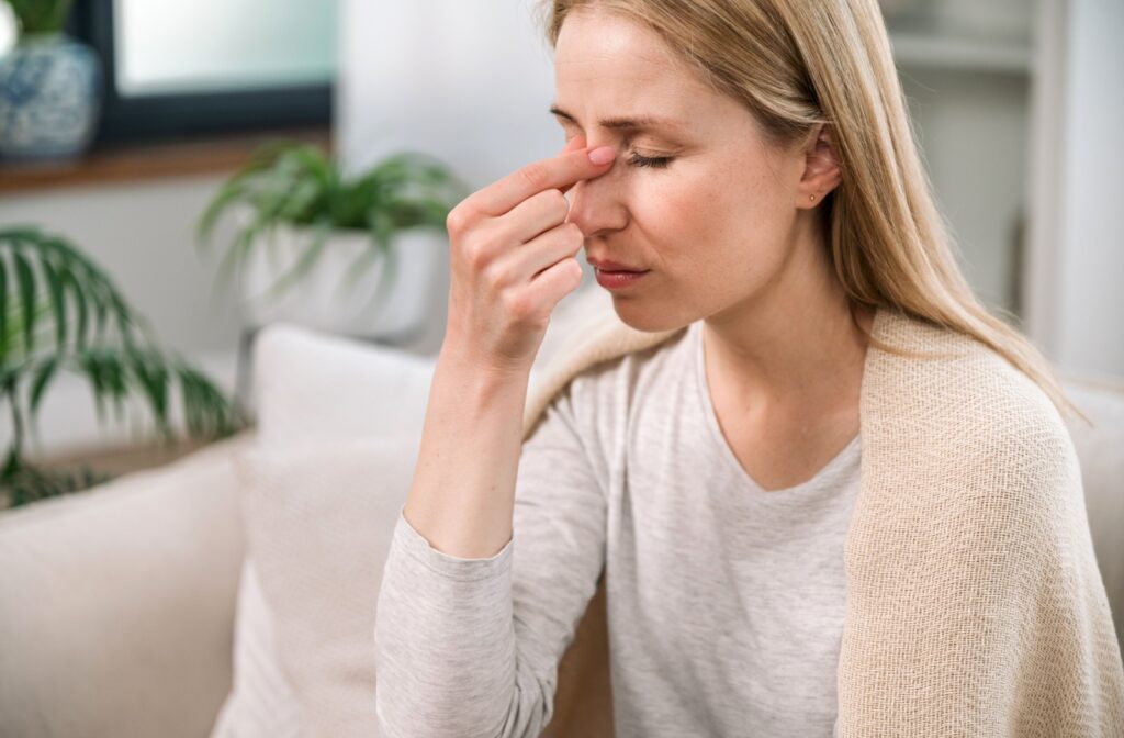 A person sitting on a couch, holding their nose due to sinus problems affecting their vision.